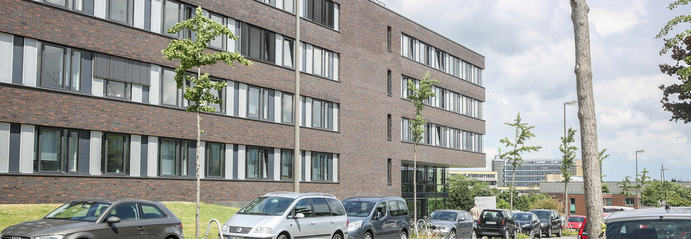 Informatik Informatikgebäude: Ein großes, braun-rot verklinkertes Gebäude mit großen Fenstern und Flachdach. Davor auf der Straße parken Autos hintereinander.